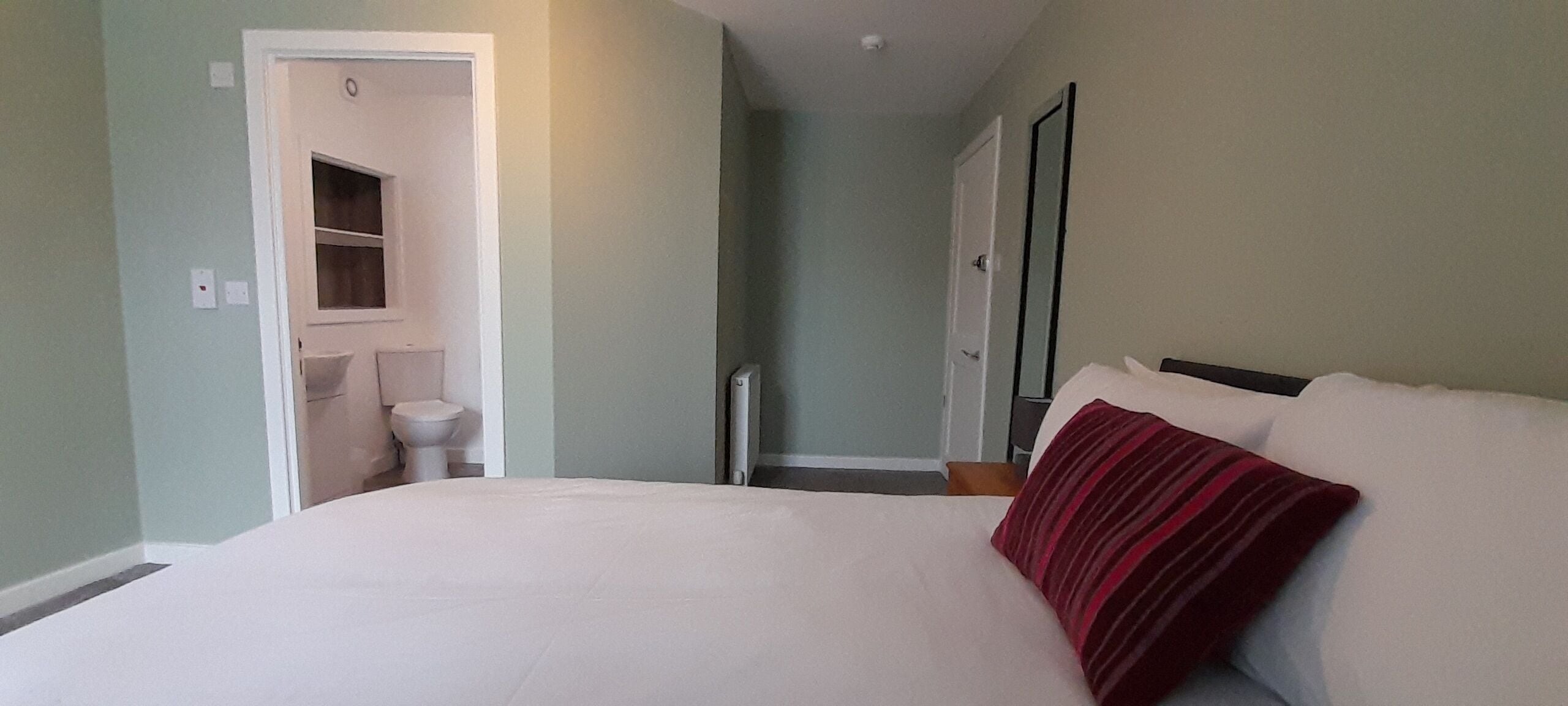A bedroom with a large bed, a red and white striped pillow, a white wall, and a door leading to a bathroom.