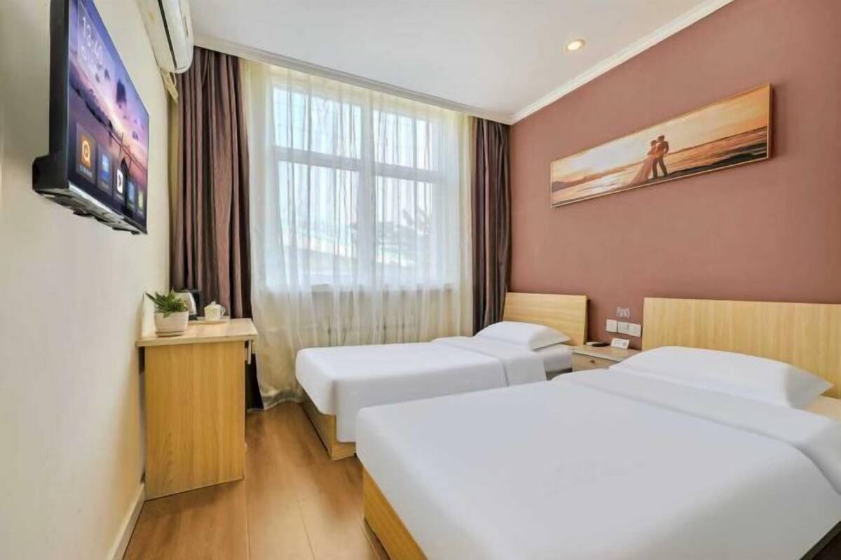 Ein Hotelzimmer mit zwei Einzelbetten, einem Schreibtisch mit Pflanze, einem Bild an der Wand und einem großen Fenster mit Vorhängen.