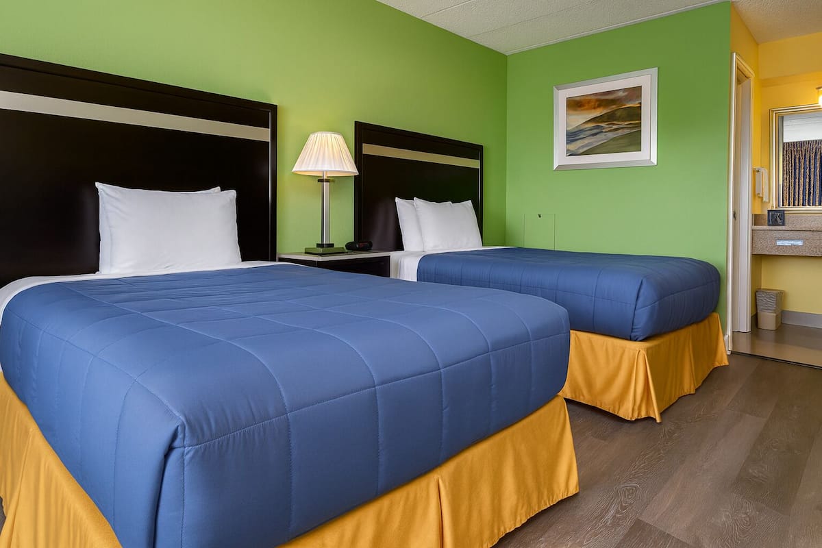 A hotel room with two beds, green walls, a painting, and a desk with a lamp.