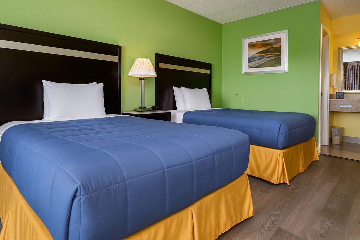 A hotel room with two beds, green walls, a painting, and a desk with a lamp.
