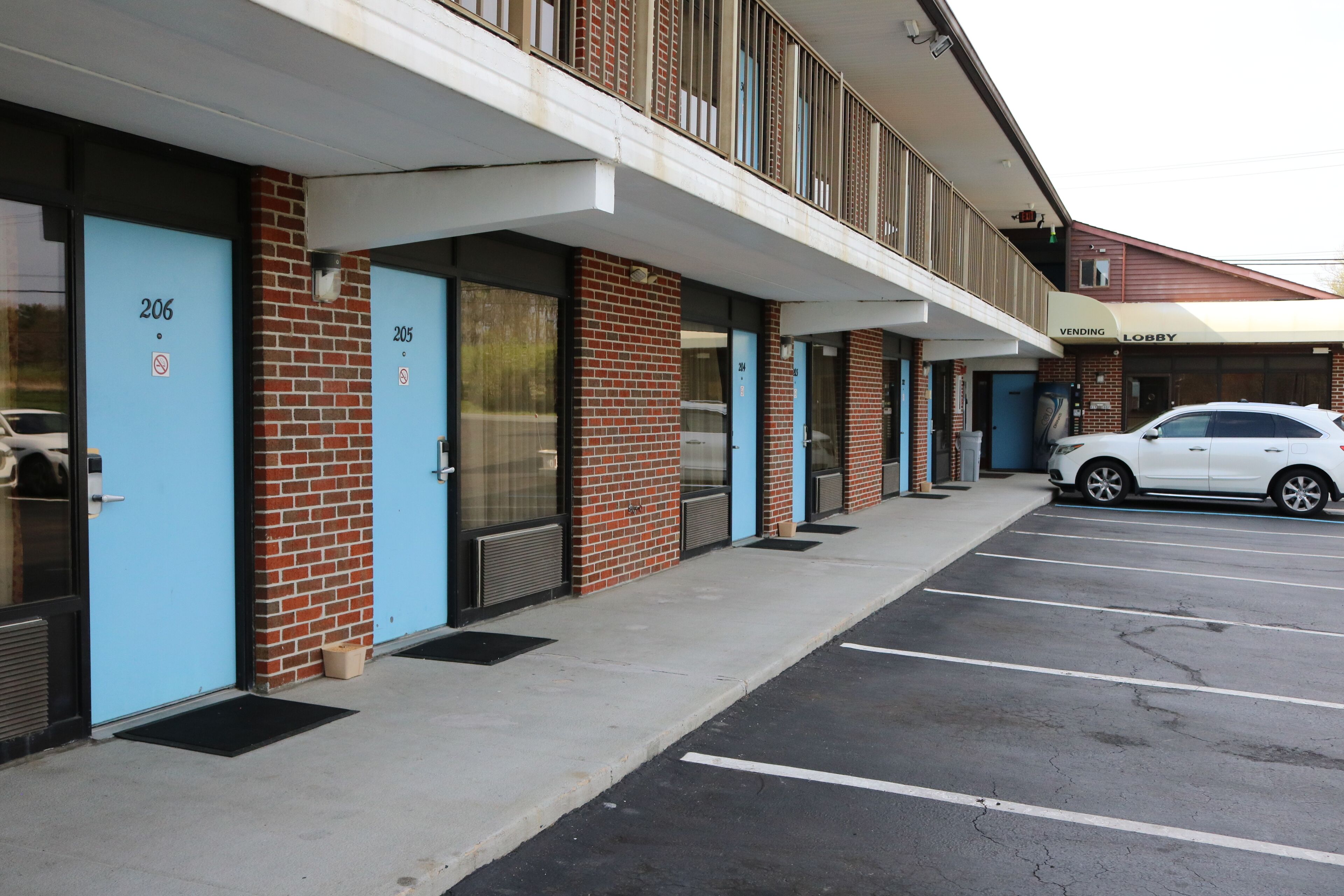 A row of blue doors with numbers 206 and 205, a white car parked in the lot, and a building with a 'Vending' and 'Lobby' sign.