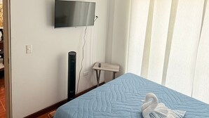 A bedroom with a blue quilted bedspread, a flat-screen TV mounted on the wall, a small bedside table, and a wall-mounted heater.