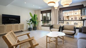 A modern living room with a large TV, comfortable seating, potted plants, and a view of a historic building through large windows.
