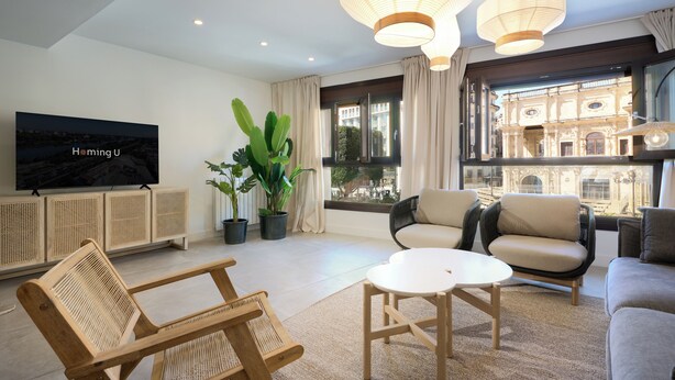 Un salon moderne doté d’un grand téléviseur, de sièges confortables, de plantes en pot et offrant une vue sur un bâtiment historique grâce à de grandes fenêtres.
