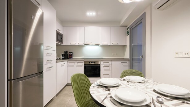 A modern kitchen with white cabinets, a stainless steel refrigerator, a microwave, and an oven. There is a dining table set with plates, glasses, and cutlery.