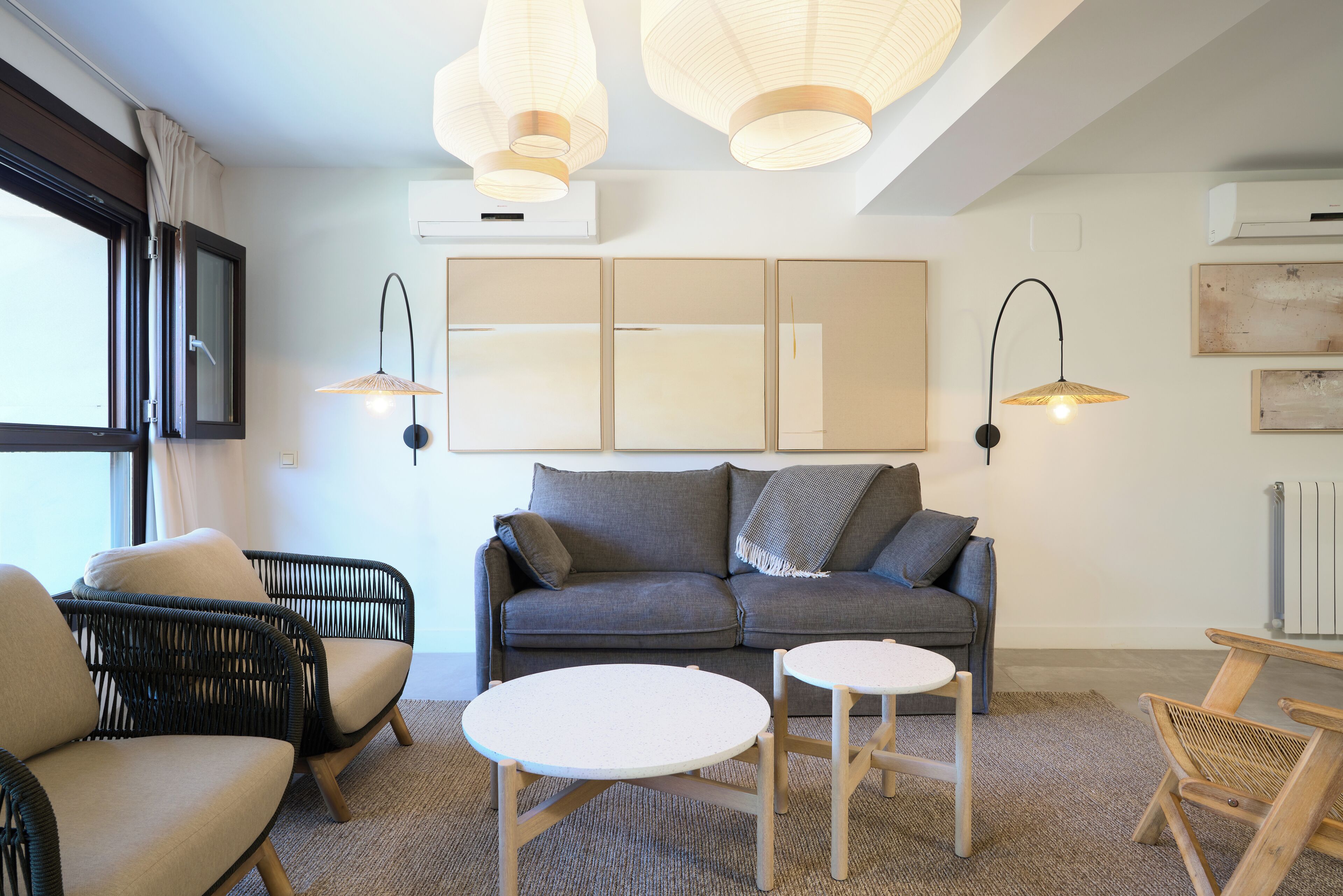 Una sala moderna con un sofá gris, dos mesas redondas, sillas de mimbre e iluminación colgante.