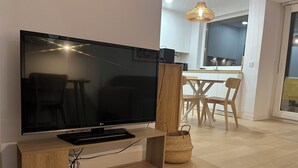 Ett modernt vardagsrum med en platt-TV på ett träställ, en matplats med trästolar och ett bord, och ett kök i bakgrunden.