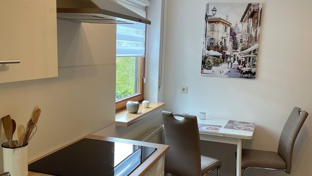 A modern kitchen with a built-in stove, a sink, and a window with blinds. There is a dining area with a table and chairs, and a painting on the wall. - Vacation apartment Philipp (Emmingen-Liptingen)