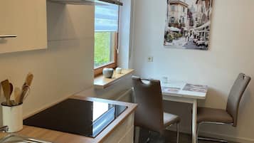 Une cuisine moderne équipée d’une cuisinière intégrée, d’un évier et d’une fenêtre avec des stores. On y trouve également un coin repas avec une table et des chaises, ainsi qu’un tableau accroché au mur.