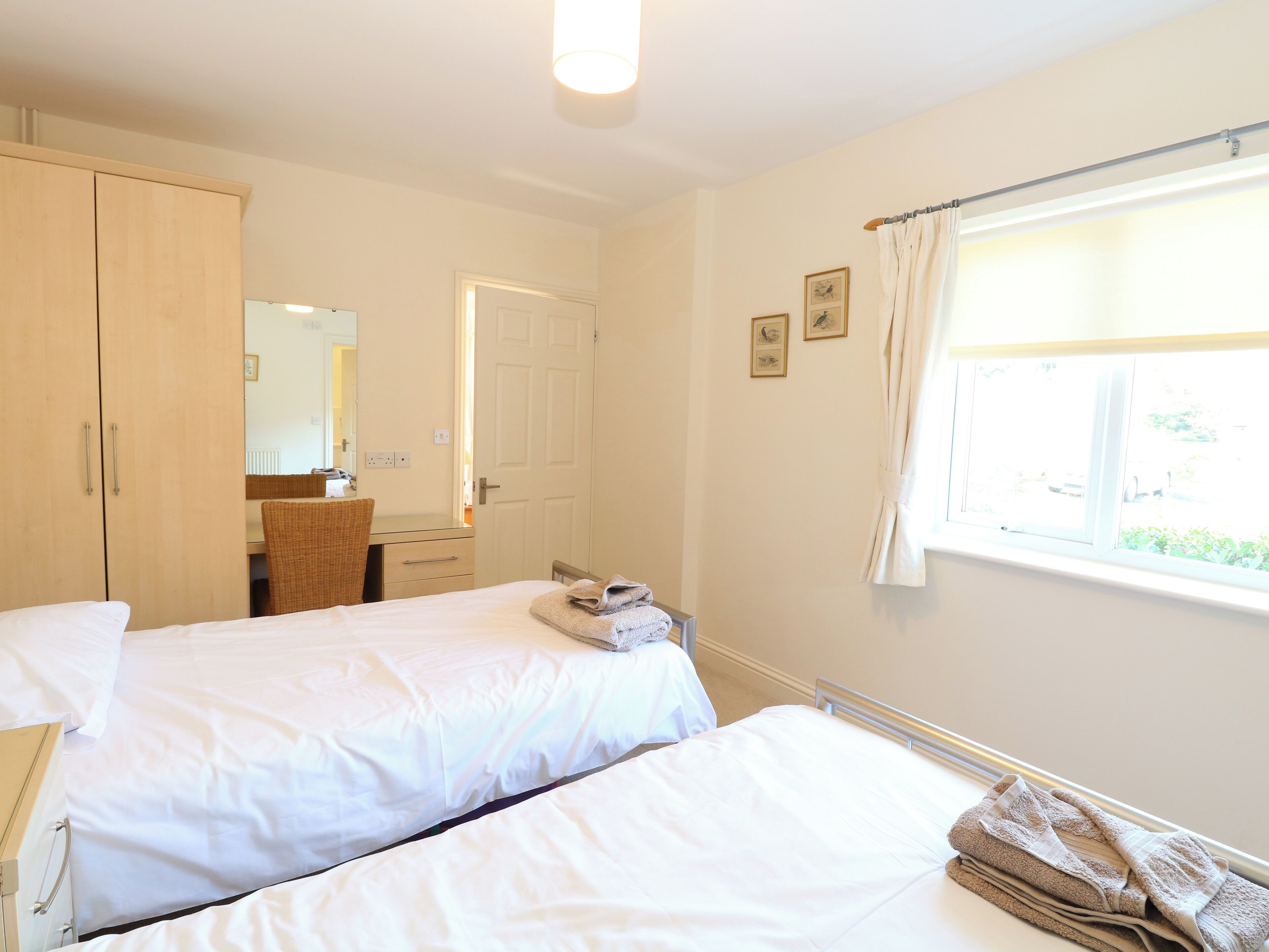 A hotel room with two single beds, a wardrobe, a mirror, a window with curtains, and a small table with a chair.