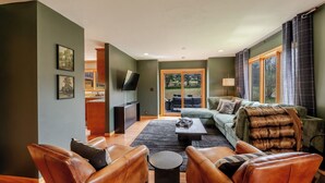 Sala de estar con sof ás de cuero, una mesa de centro y un televisor. Se aprecia una ventana grande con cortinas y vistas a árboles.