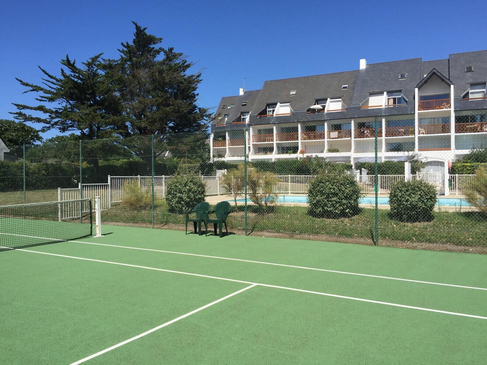 Uma quadra de tênis com uma rede, cadeiras verdes e uma piscina ao fundo.