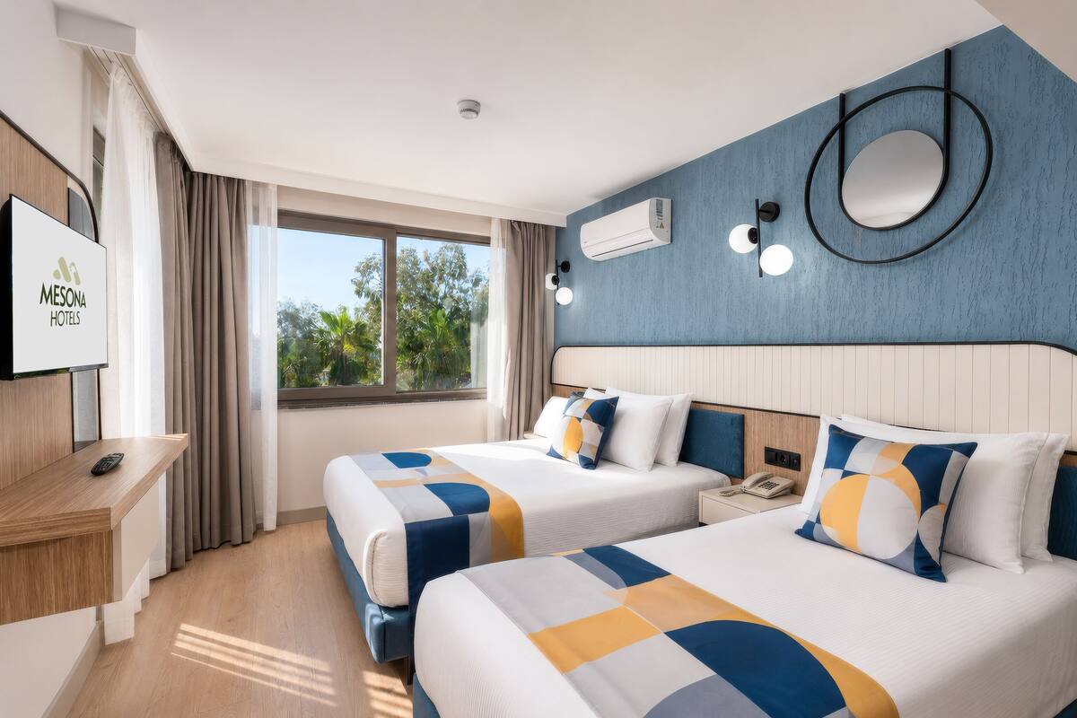 Une chambre d’hôtel avec deux lits, une télévision, un bureau et une fenêtre donnant sur des arbres.