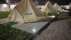A row of beige tents labeled "VEVOR" on a concrete platform with a green mat in front, set against a night sky with a building in the background. - Glamphouse, adventure and relaxation! (Oaxtepec)