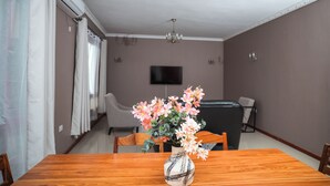 Un comedor con una mesa de madera, sillas, un jarrón con flores, un televisor en la pared y un sofá al fondo.