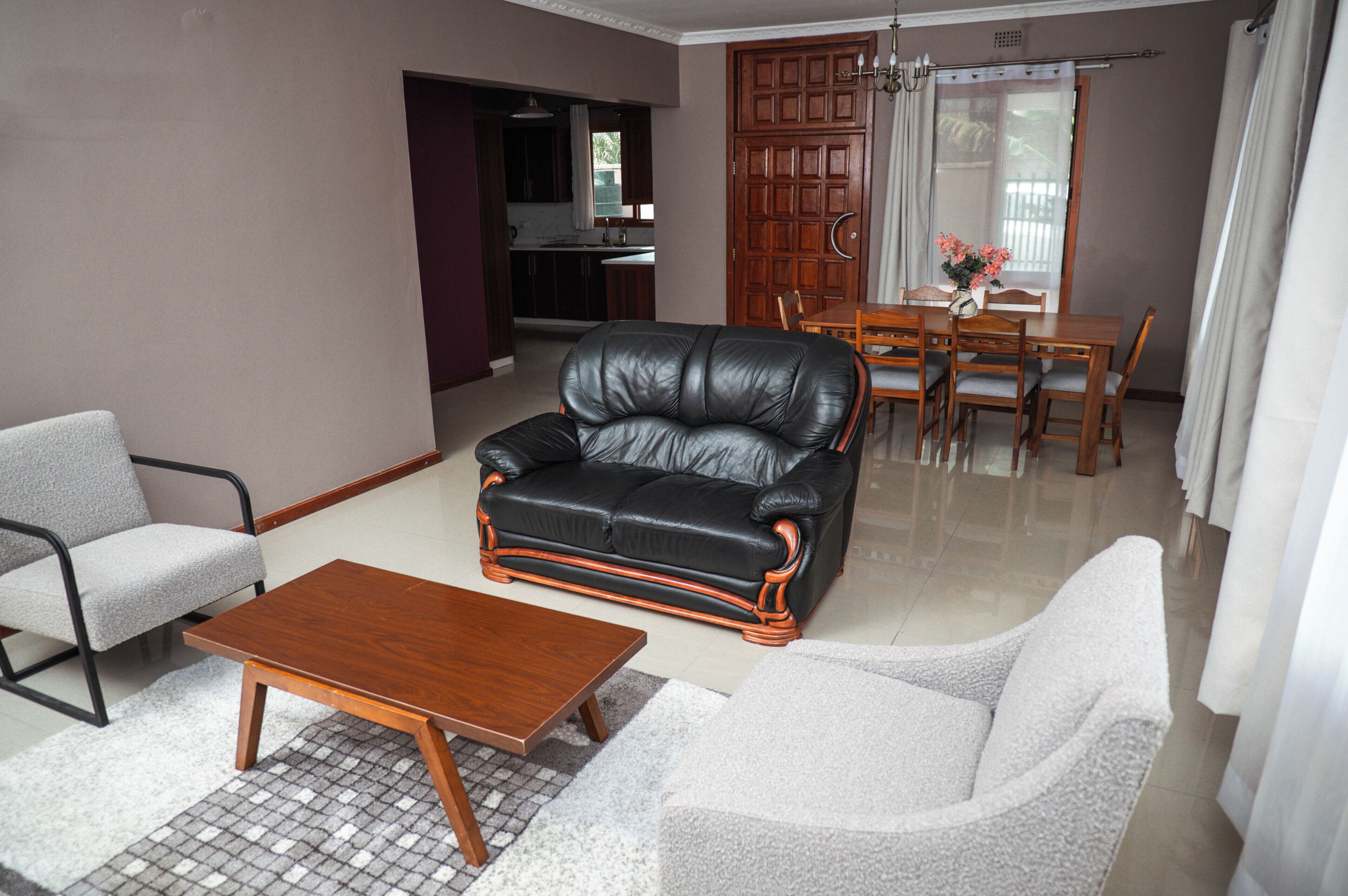 A living room with a black leather sofa, a wooden coffee table, and a grey armchair. There is a dining area with a wooden table and chairs.
