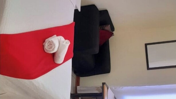 A hotel room with a white bed, red pillows, a black chair, and a desk with a lamp. - Marcifen (Harare)