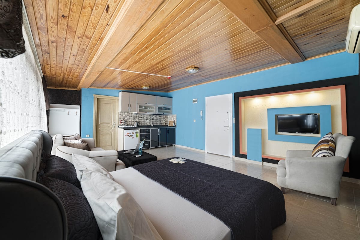 A modern room with a wooden ceiling, a bed, a sofa, a TV, and a kitchen area in the background.