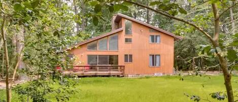 A modern wooden house with large windows, surrounded by a grassy yard and trees.