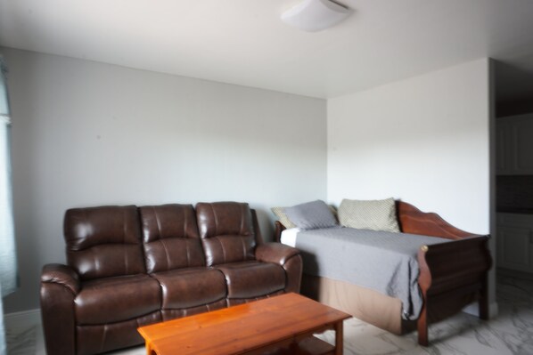 A living room with a brown leather sofa, a wooden coffee table, and a bed with a grey blanket. - Mary Pop Apartments (Dania Beach)
