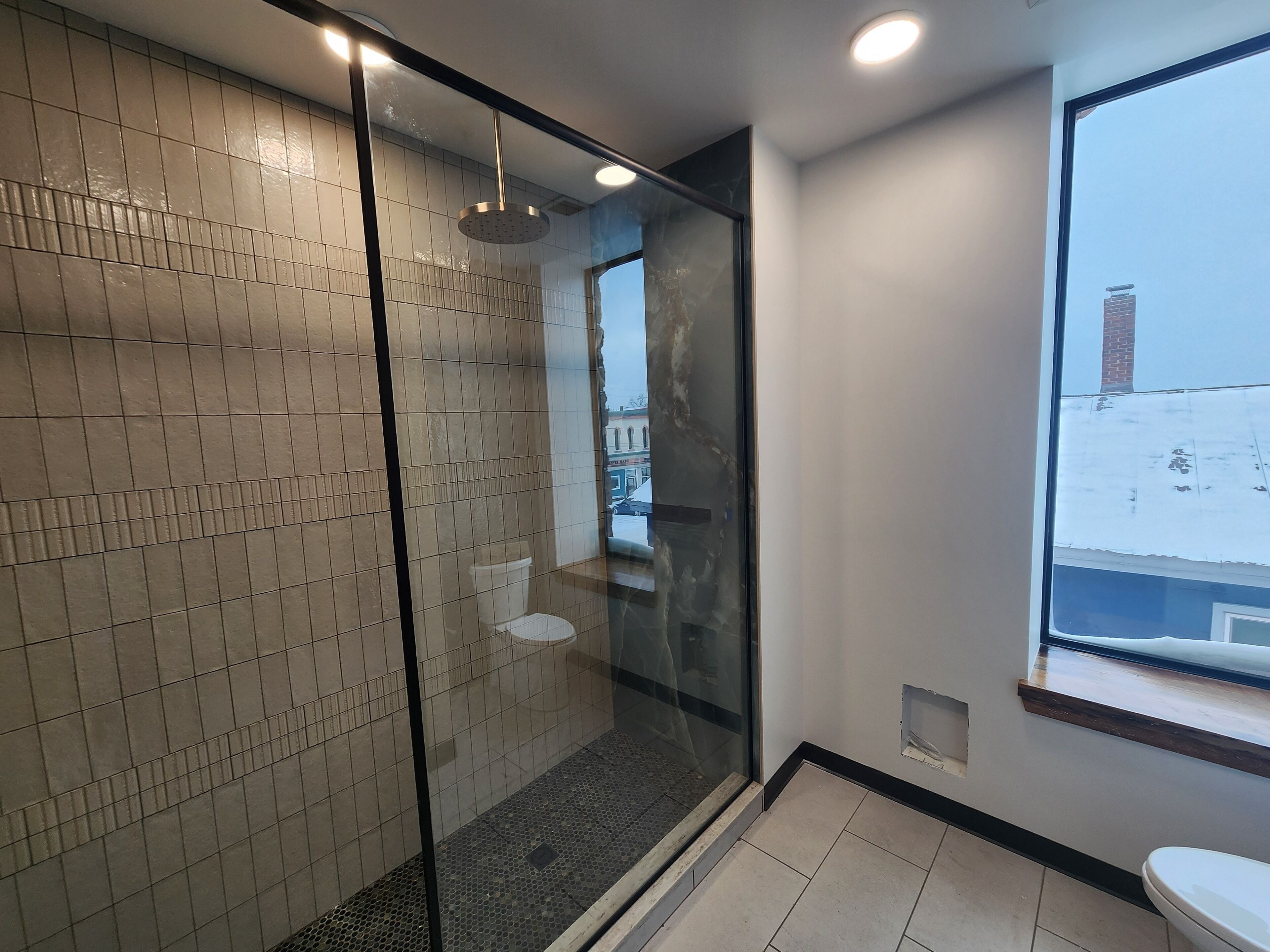 Une salle de bain moderne avec une douche à l’italienne en verre, des murs carrelés et une grande fenêtre donnant sur l’extérieur.