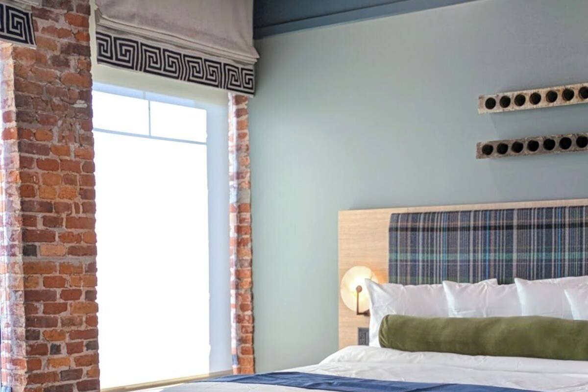 Habitación de hotel con pared de ladrillo, una cama grande con ropa blanca, un banco y una ventana con cortinas.