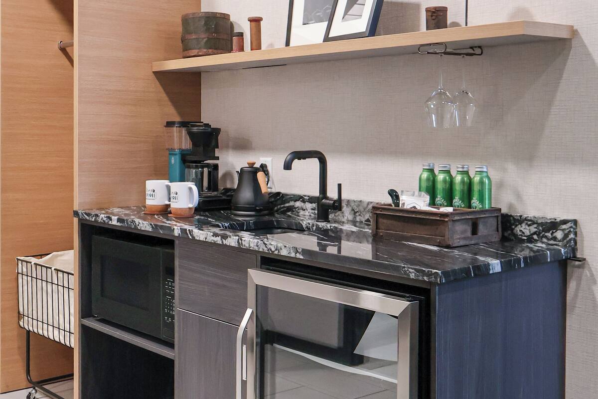 Cocina compacta con microondas, horno y refrigerador integrados. Hay un estante de madera sobre el lavamanos con una tetera, una cafetera y algunas botellas.