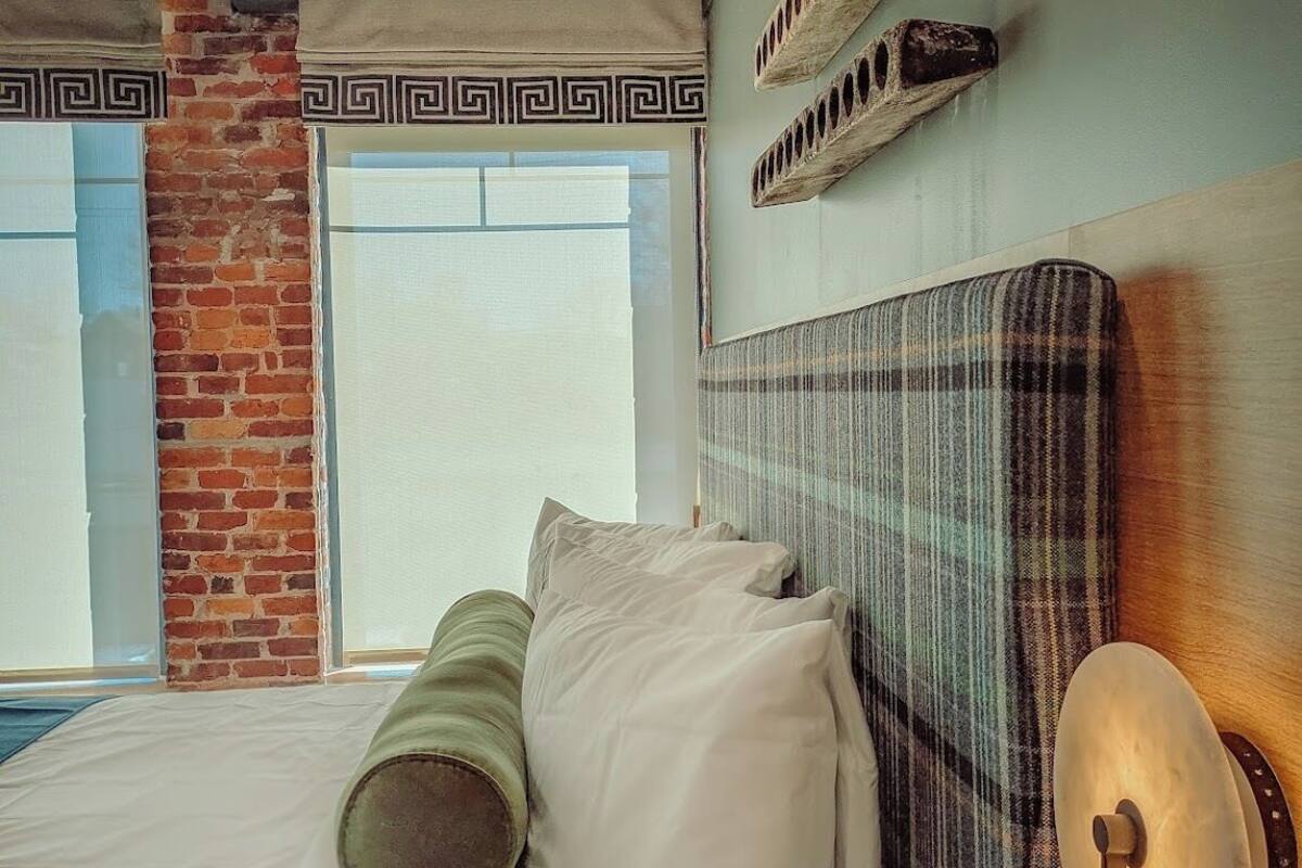 Habitación de hotel con pared de ladrillo, una cama grande con ropa blanca, cabecero con reloj y ventana con vistas.