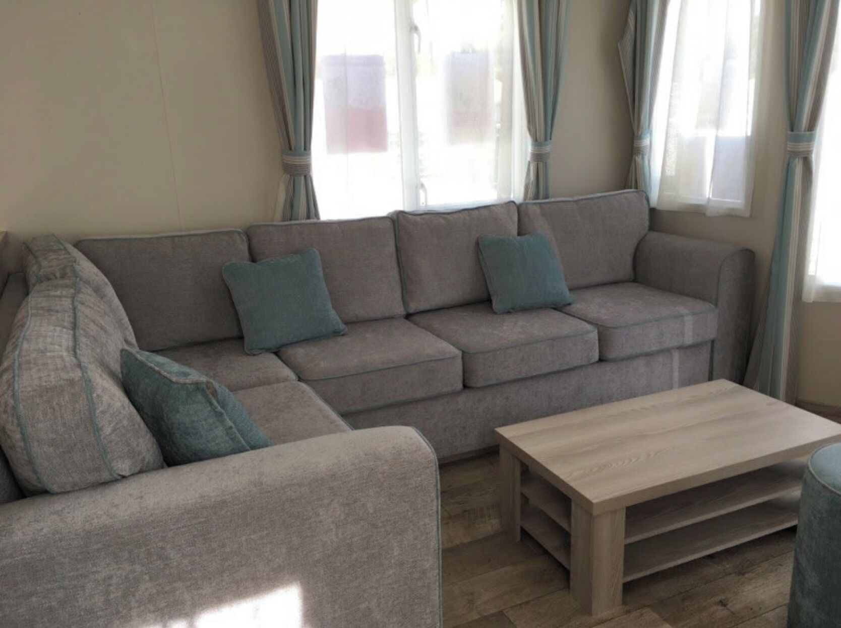 Un salon moderne avec un canapé d’angle gris, deux fauteuils assortis, une table basse en bois et des rideaux bleu clair.