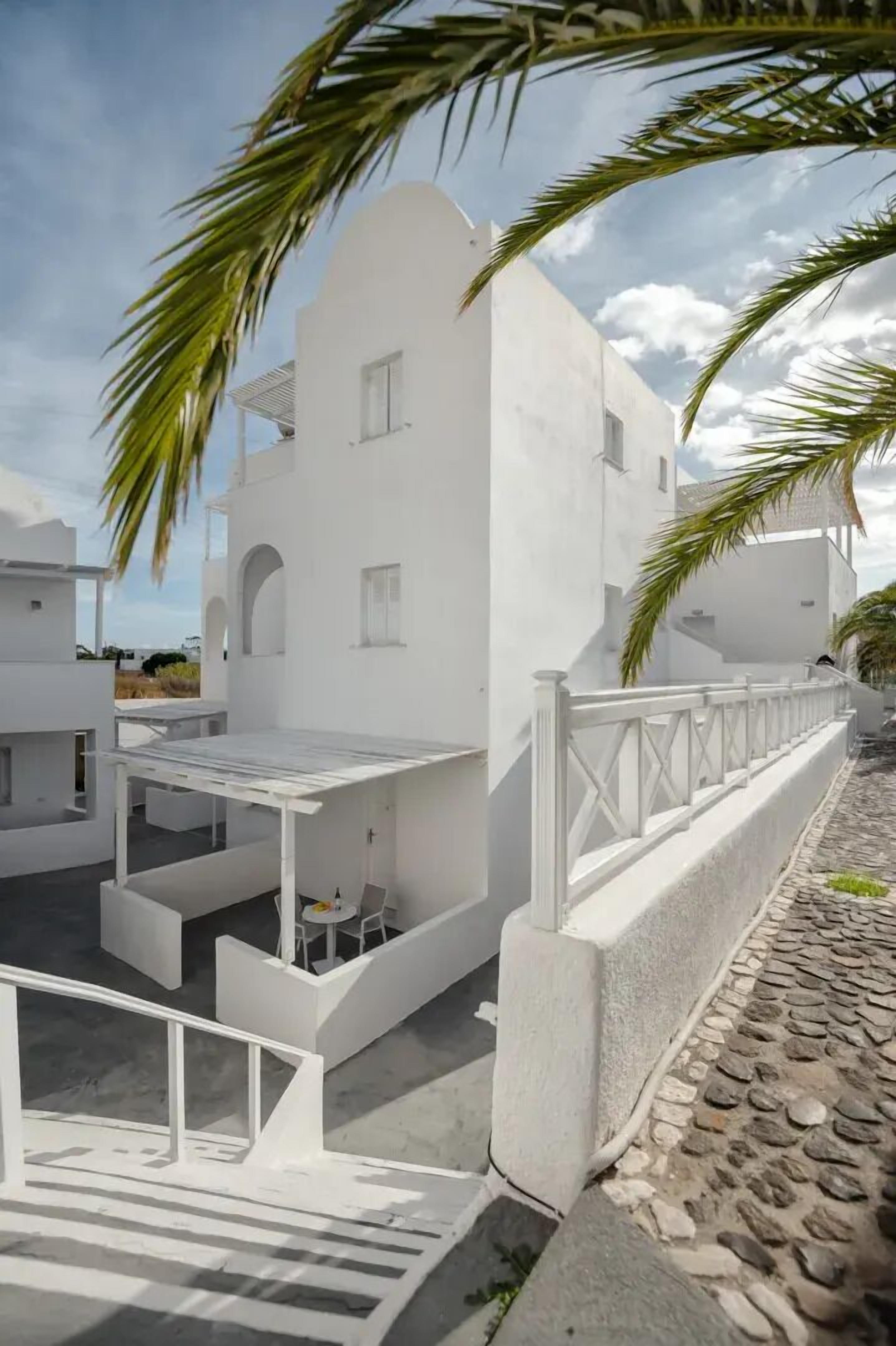 Um edifício branco com área de estar coberta ao ar livre, um caminho pavimentado em pedra e uma palmeira.