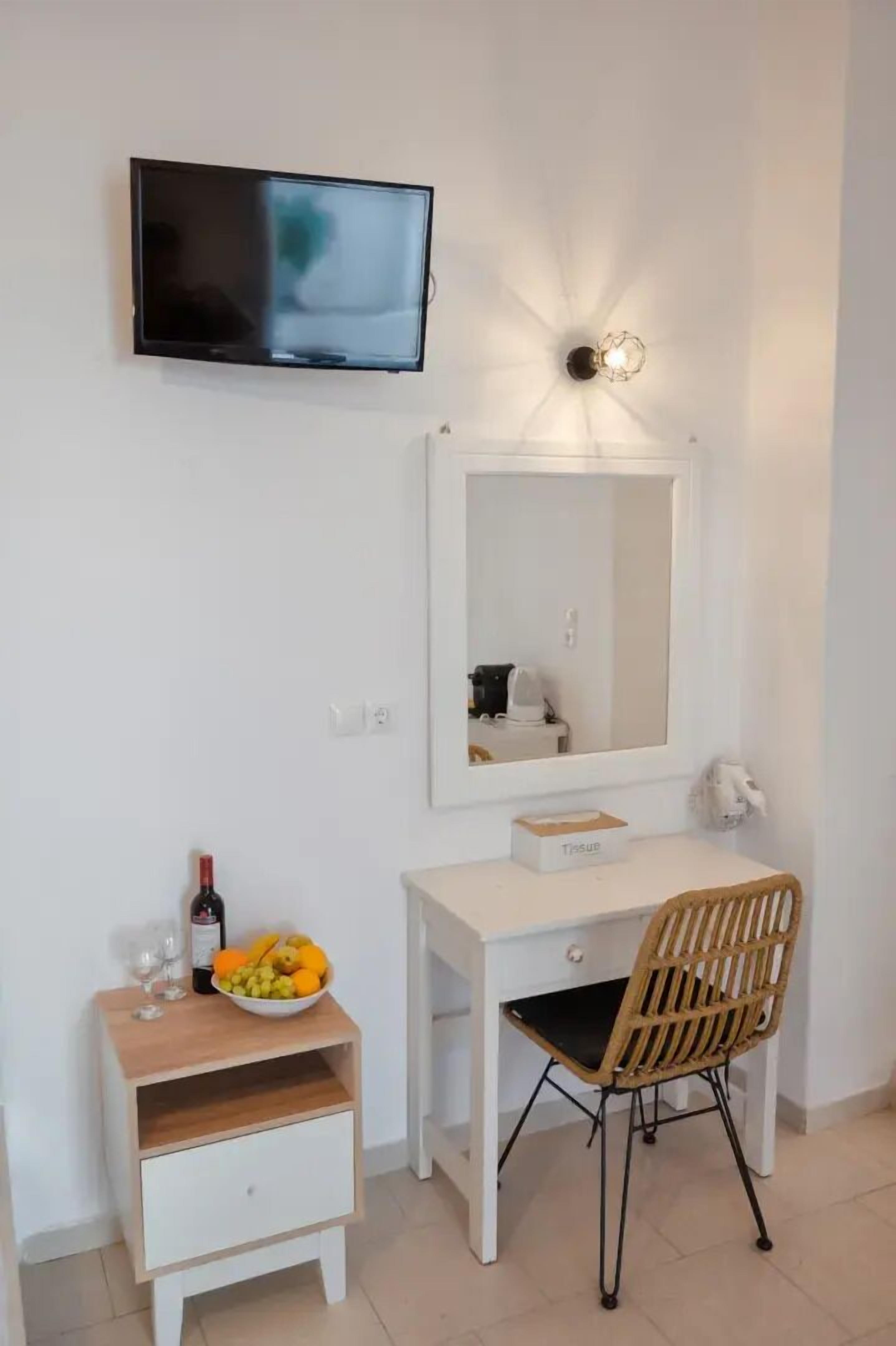 Una habitación pequeña con un televisor montado en la pared, un espejo sobre el escritorio, una silla de madera, una mesita de noche con un frutero y copas de vino, y un escritorio blanco.