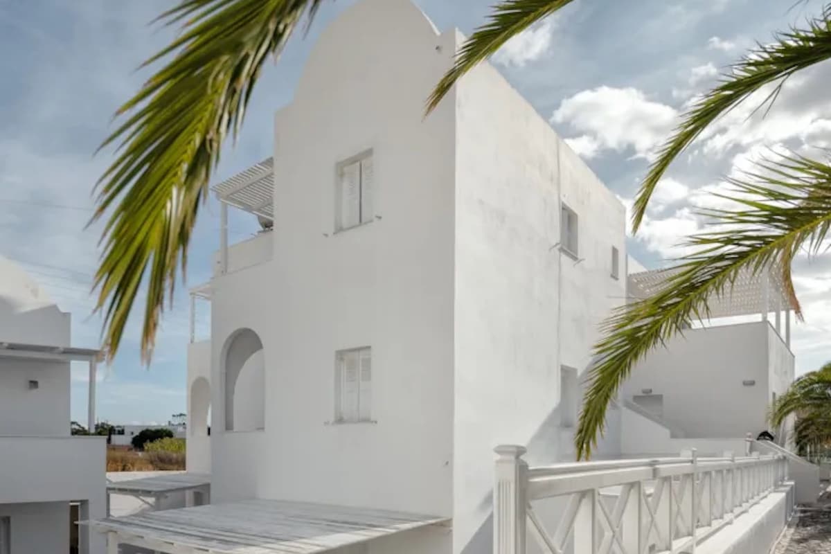 A white building with a covered outdoor seating area, a stone pathway, and a palm tree.