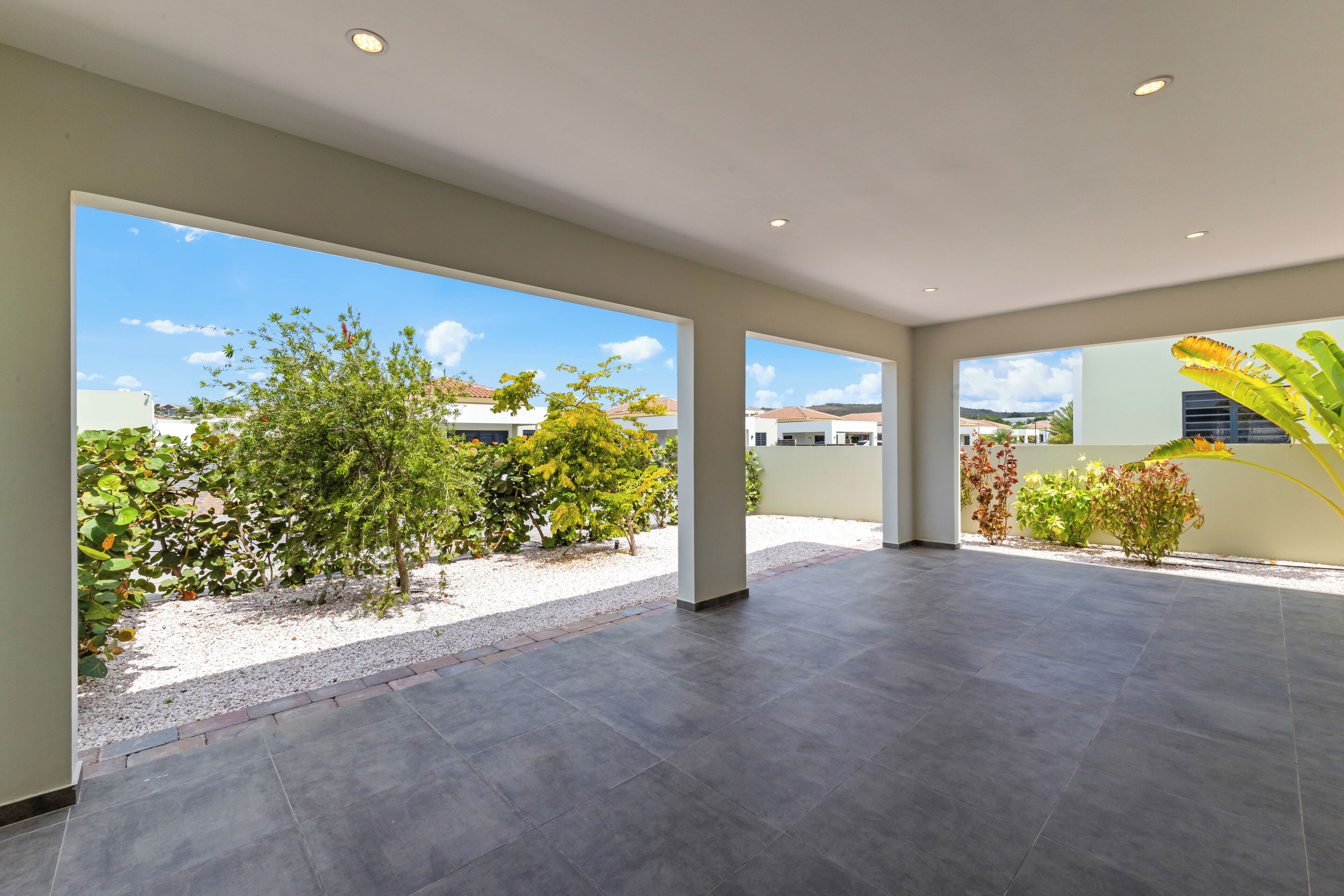 A spacious indoor area with large windows, a tiled floor, and a view of a garden and buildings outside.