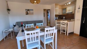 Un coin repas avec une table et des chaises blanches, une bouteille de vin et un saladier de fruits.