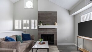Ein modernes Wohnzimmer mit einem grauen Sofa, einem weißen Couchtisch, einem Kamin und einem Fernseher auf einem Holzgestell.