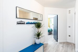 Um corredor moderno com parede branca, um aparador azul, uma planta em vaso, um espelho e uma porta que dá para um quarto.