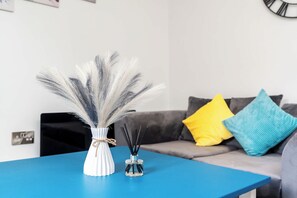Quarto com uma mesa azul, um vaso branco com penas e uma garrafa de vidro com gravetos pretos. Há um sofá cinza com almofadas amarelas e azuis.