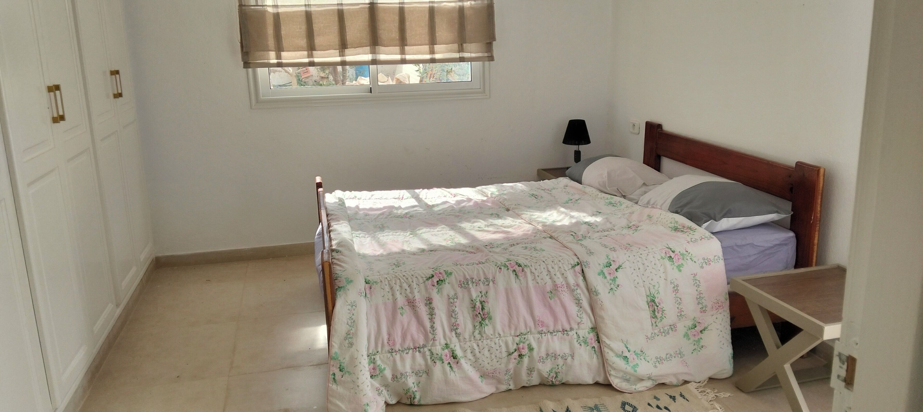 Une chambre à coucher avec un lit, une table de chevet, une fenêtre avec des stores et une porte dotée d’une poignée.