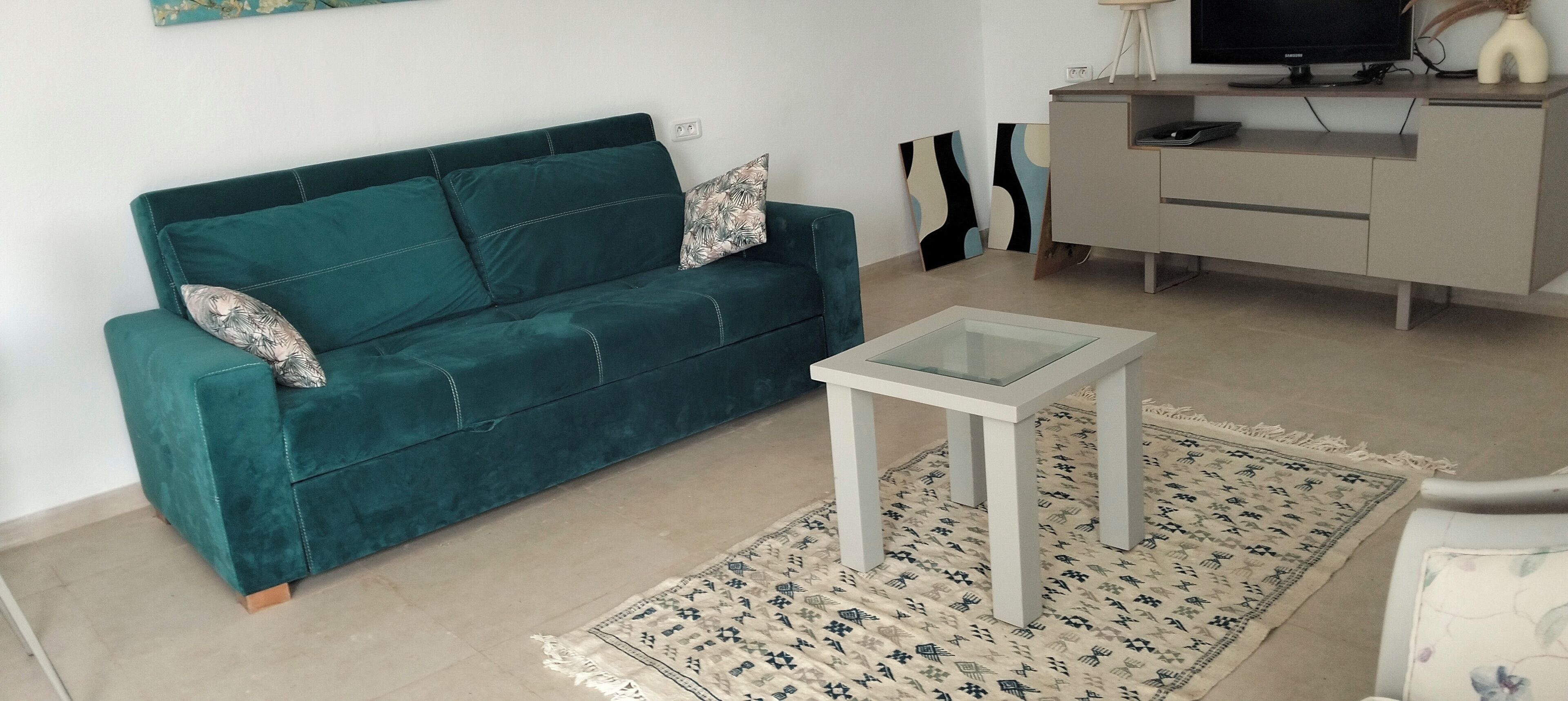 A teal sofa with patterned cushions, a small white table, and a patterned rug in a living area.