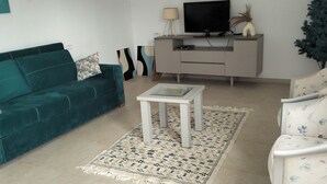 A teal sofa, a white coffee table, a floral armchair, a TV on a cabinet, and a small plant. - Quiet house with garden Pets welcome (Sakiet Ezzit)