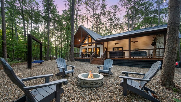 Una cabaña con chimenea de piedra rodeada de sillas Adirondack, ubicada en un bosque.