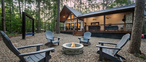 Una cabaña con chimenea de piedra rodeada de sillas Adirondack, ubicada en un bosque.