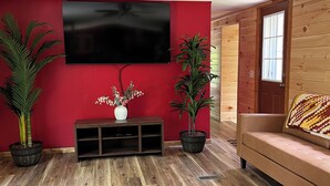 A living room with a red wall, wooden flooring, a ceiling fan, a flat-screen TV, two potted plants, a dark leather sofa, and a wooden cabinet. - White Pines B&B LLC (Blackduck)