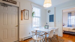 A room with a white door labeled 'FARMHOUSE,' a dining table with chairs, a hanging light fixture, a window with a plant, and a bed with patterned curtains. - Amazing West End apartment walkable to downtown and the waterfront. (Portland)