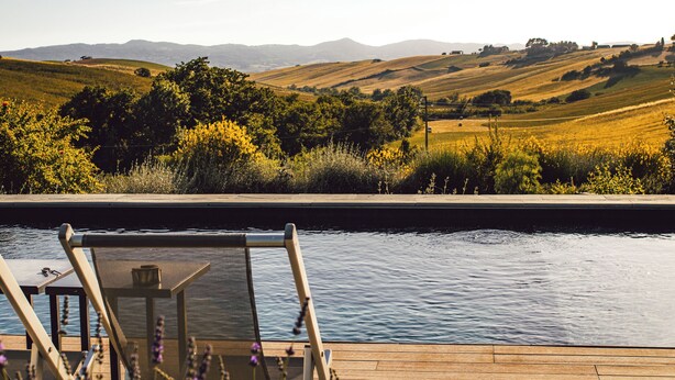 A poolside view with a wooden deck, a table with glasses, and a view of rolling hills and mountains in the background. - Boutique eco Agriturismo nestled among ancient olive trees and stunning Tuscan hills (Santa Luce)