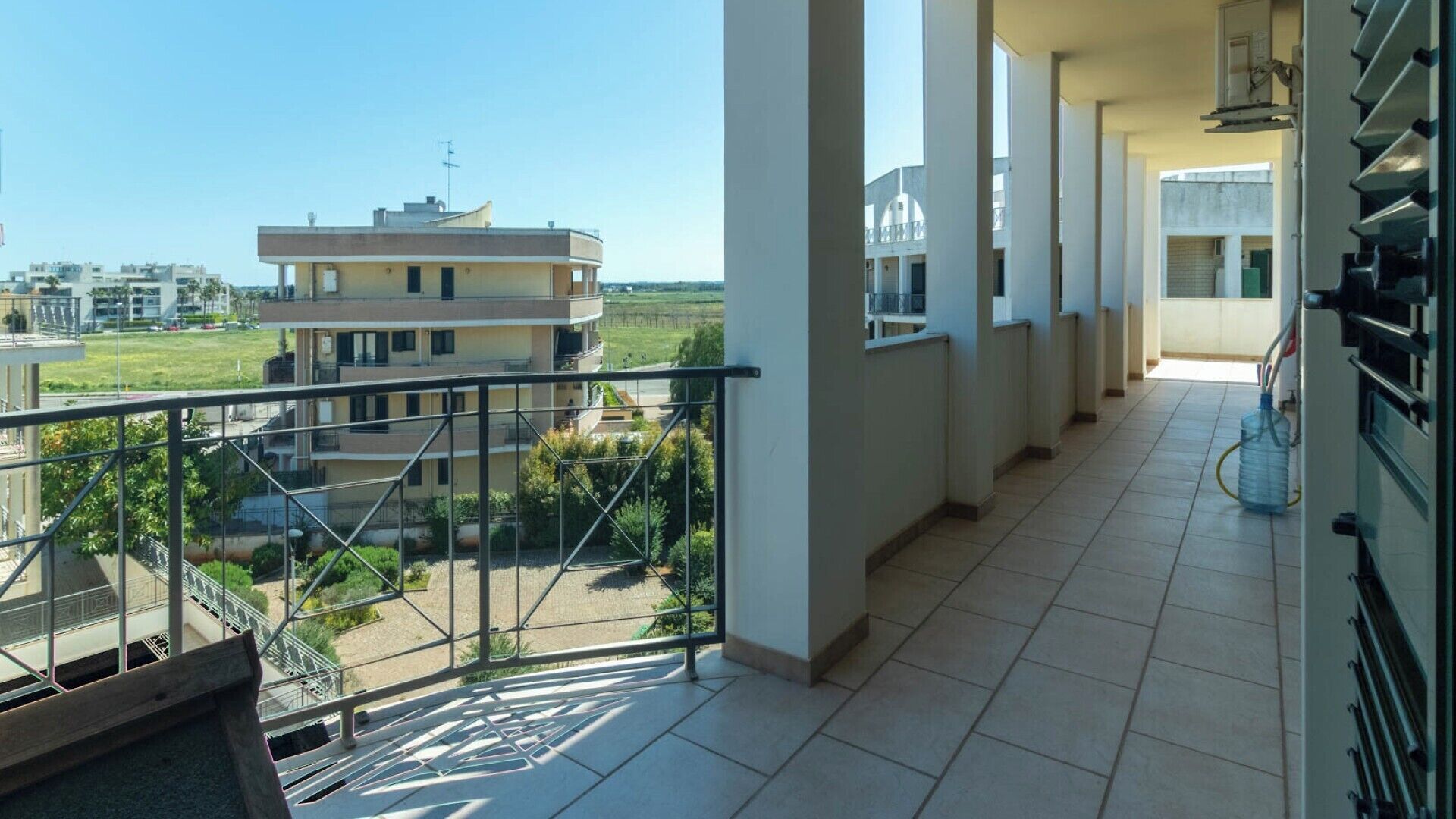 Un balcón con vistas a una zona residencial, suelo de baldosas y un dispensador de agua.