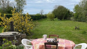A round table with a checkered tablecloth, two white plastic chairs, and a wooden chair. The table is set with a white mug, a white pitcher, and a tray with cups and a teapot. The setting is outdoors with a grassy area and trees in the background. - A unique stay in an old railway station, in the heart of the Cotentin sea & countryside. (Saint-Georges-de-la-Rivière)