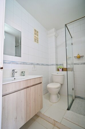 Un baño con lavamanos, espejo e inodoro blancos, cabina de ducha con mampara de vidrio y suelo de baldosas.