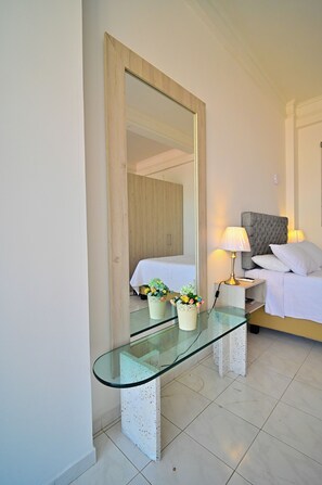 Habitación de hotel con escritorio de vidrio, cama con ropa blanca, cabecera gris y lámpara de noche.