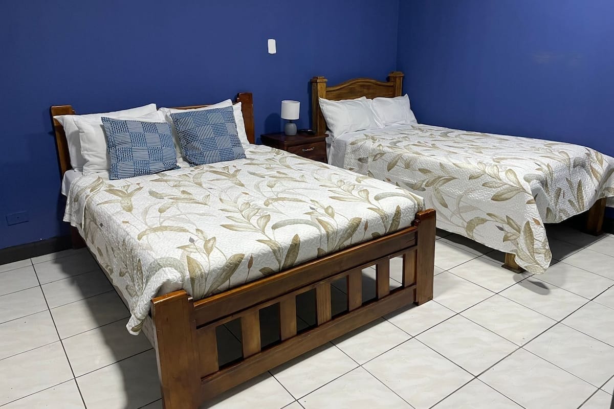 Duas camas de solteiro com estrutura de madeira e roupa de cama floral, um criado-mudo com abajur, e uma parede azul.